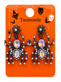 A pair of black spider drop earrings with iridescent purple and blue rhinestones, displayed on an orange Taramanda brand Halloween card.