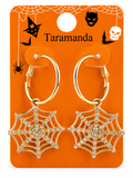 A pair of gold hoop earrings with dangling spiderweb charms embellished with clear rhinestones, displayed on an orange Halloween-themed card labeled 'Taramanda'.
