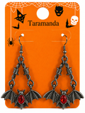 A pair of gothic bat drop earrings with silver-toned metal chains, bat wings, and red oval gemstones, displayed on an orange Halloween-themed card labeled 'Taramanda'.