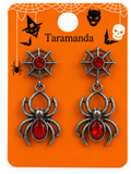 A pair of gothic spider drop earrings with antique silver metal spider bodies and red faceted gemstones, displayed on an orange Halloween-themed backing card labeled 'Taramanda'.