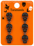 A pair of gothic skull drop earrings on an orange Halloween-themed display card. The earrings feature three stacked skull charms in a blackened silver metal setting, embellished with small black rhinestones and leaving dark red-brown recesses.