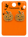 A pair of Halloween pumpkin stud earrings with gold-tone rhinestone detailing and green stems, displayed on an orange card labeled 'Taramanda' with spiderweb and Halloween graphics.