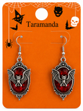 A pair of gothic-style bat drop earrings with red faceted gemstones and silver-toned metal frames, displayed on an orange Halloween-themed backing card labeled 'Taramanda'.