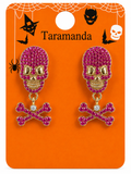 A pair of Halloween-themed dangle earrings on a branded orange backing card. The earrings feature skull tops paved with bright pink rhinestones with gold-tone eyes and mouth details, hanging from gold-tone rings above matching pink rhinestone-crossbones.
