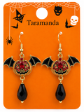 A pair of Halloween-themed bat drop earrings on an orange display card. The earrings feature gold-tone metal, black enamel wings and teardrop beads, and red crystal accents around a central bat-shaped motif.