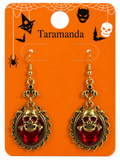 A pair of gothic-style dangle earrings with red skull and crossbones rhinestones in ornate gold frames, displayed on an orange Halloween-themed card labeled 'Taramanda'.
