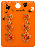 A pair of Halloween-themed drop earrings featuring three stacked pumpkin charms with carved jack-o'-lantern faces, displayed on an orange card labeled 'Taramanda' with spiderwebs, bats, and a skull.