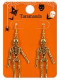 A pair of gold skeleton drop earrings displayed on an orange Halloween-themed backing card labeled 'Taramanda' with spiderwebs, bats, a witch hat, and a skull.