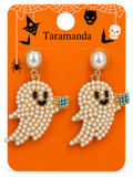 A pair of Halloween ghost dangle earrings with cream pearl-like beads, gold-tone metal, black embroidered eyes and mouth, and turquoise blue beaded accents on the sleeves, displayed on an orange card labeled Taramanda with spiderweb and Halloween graphics.
