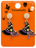 A pair of Halloween-themed witch hat dangle earrings on a branded orange display card. The earrings have black witch hats with purple bands, gold stars, white faux pearl studs, and purple bows.