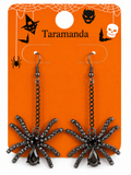 A pair of black spider drop earrings with rhinestone-embellished legs and teardrop crystal pendants, displayed on an orange Halloween-themed card labeled 'Taramanda'.