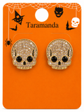 A pair of gold skull stud earrings covered in clear rhinestones with black gemstone eyes, displayed on an orange Halloween-themed card labeled Taramanda.
