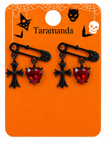 A pair of gothic-style safety pin earrings with black crosses and red heart-shaped rhinestones, displayed on an orange Halloween-themed card labeled 'Taramanda'.