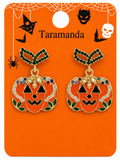 A pair of Halloween-themed pumpkin earrings on a branded orange display card. The earrings have gold-tone metal outlines, orange enamel, black facial features, green leaf accents, and rhinestone borders with small blue and purple stones.